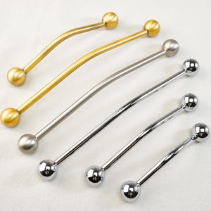 Pode incluir: Uma variedade de puxadores de armário em acabamentos dourados e prateados. Cada puxador apresenta uma barra curva com uma extremidade esférica em cada lado. Os puxadores estão dispostos sobre uma superfície branca, mostrando seus diferentes tons metálicos e designs.