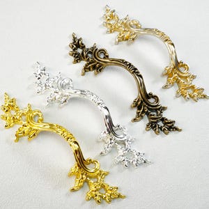 May include: Four decorative cabinet handles in gold, silver, and bronze tones. Each handle features an ornate, curved design with floral accents. The handles are arranged on a white surface, showcasing their intricate details.