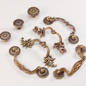 May include: An assortment of antique brass cabinet hardware, including knobs and handles. The knobs feature a floral design, while the handles have ornate, curved shapes. The hardware is arranged on a white surface, showcasing the details and finish.