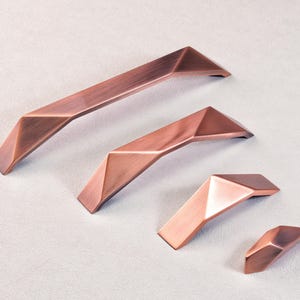 Könnte beinhalten: Kupferfarbenes Möbelbeschlagsset. Das Set enthält zwei lange Griffe, einen mittleren Griff und einen kleinen Knauf. Jedes Teil hat ein geometrisches, facettiertes Design und eine warme, metallische Oberfläche.