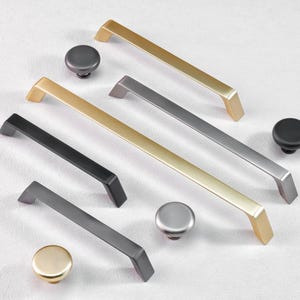 May include: A collection of cabinet hardware, including gold, black, and silver-toned handles and knobs. The handles are rectangular with angled ends, and the knobs are round. The hardware is displayed on a white surface.