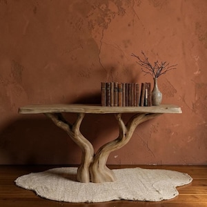 May include: A natural wood console table with a tree trunk base. The table supports a collection of antique books and a small vase with branches. The table sits on a cream-colored rug against a reddish-brown wall.
