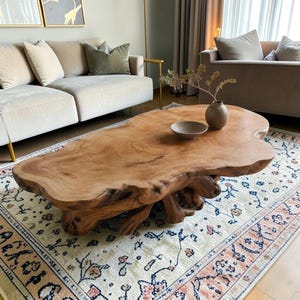 May include: A large, natural wood coffee table with an organic shape. The light brown table has visible wood grain and sits on a base of intertwined wood. A small, brown vase and bowl are on the table. The table is on a patterned rug.