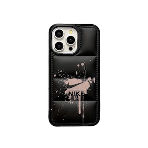 May include: Black phone case with a quilted design and a pink Nike logo. The case has a splatter paint effect and the word "NIKE" printed below the logo. The case is designed to fit a smartphone.