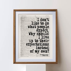 May include: A framed print with text on a vintage dictionary page. The black text reads, "I don't like to do what people expect. Why should I live up to their expectations instead of my own?" The frame is brown.