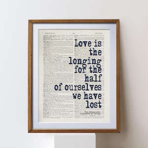 May include: A framed print with a quote on a vintage book page. The dark blue text reads "Love is the longing for the half of ourselves we have lost". The frame is a brown colour.