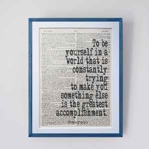 May include: Framed print with a blue wooden frame and white matting. The print features a vintage dictionary page with a quote in bold black text: "To be yourself in a world that is constantly trying to make you something else is the greatest accomplishment."
