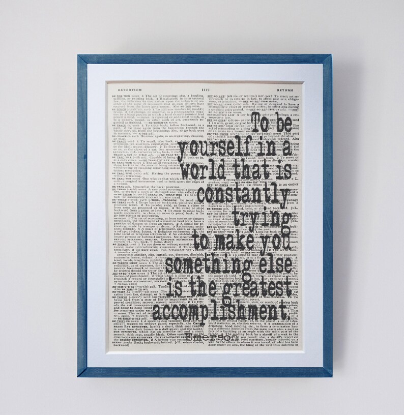 May include: Framed print with a blue wooden frame and white matting. The print features a vintage dictionary page with a quote in bold black text: "To be yourself in a world that is constantly trying to make you something else is the greatest accomplishment."
