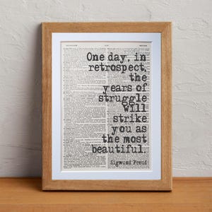 May include: A framed print featuring a quote by Sigmund Freud: "One day, in retrospect, the years of struggle will strike you as the most beautiful." The text is in black, set against a vintage book page background, and framed in light brown wood.