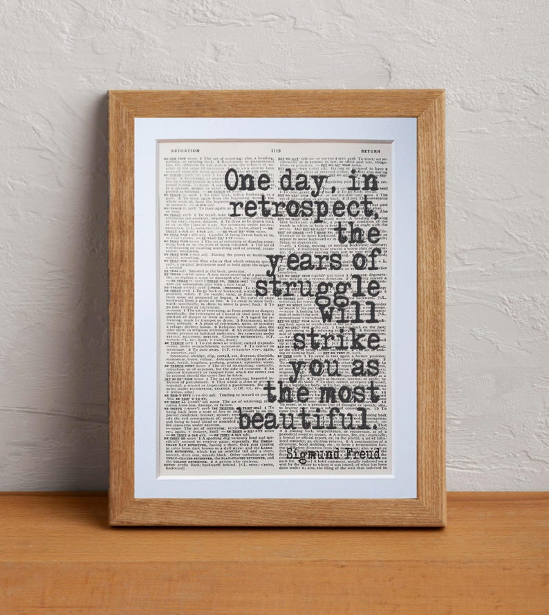 May include: A framed print featuring a quote by Sigmund Freud: "One day, in retrospect, the years of struggle will strike you as the most beautiful." The text is in black, set against a vintage book page background, and framed in light brown wood.
