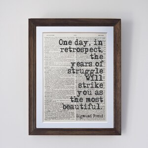 May include: Framed print featuring a vintage newspaper background with the quote "One day, in retrospect, the years of struggle will strike you as the most beautiful" by Sigmund Freud. The frame is dark brown.