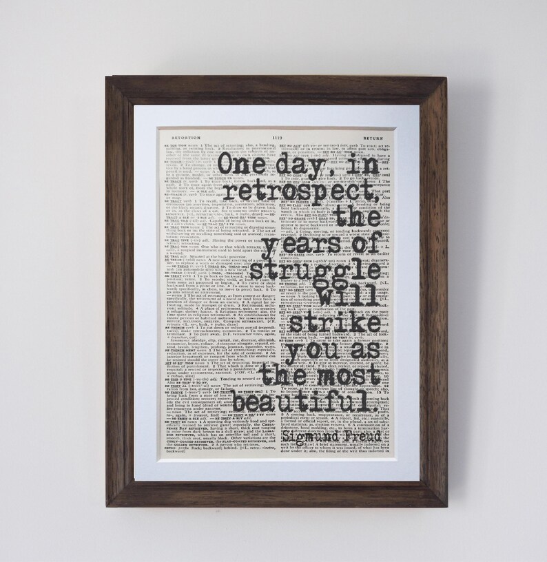 May include: Framed print featuring a vintage newspaper background with the quote "One day, in retrospect, the years of struggle will strike you as the most beautiful" by Sigmund Freud. The frame is dark brown.