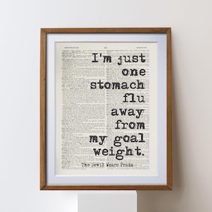 May include: A framed print with a brown wooden frame. The print features black text on a vintage dictionary page. The text reads: "I'm just one stomach flu away from my goal weight." The Devil Wears Prada is at the bottom.