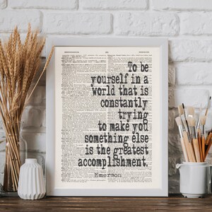 May include: Framed print with the quote "To be yourself in a world that is constantly trying to make you something else is the greatest accomplishment." The text is in black on a vintage book page.