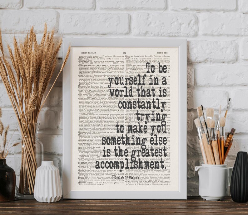 May include: Framed print with the quote "To be yourself in a world that is constantly trying to make you something else is the greatest accomplishment." The text is in black on a vintage book page.