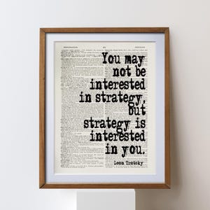 May include: Framed print featuring a quote by Leon Trotsky: "You may not be interested in strategy, but strategy is interested in you." The text is in black, set against a vintage dictionary page background, within a brown wooden frame.