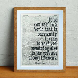 May include: A framed print with a quote on a vintage dictionary page. The quote reads, "To be yourself in a world that is constantly trying to make you something else is the greatest accomplishment." The frame is a light brown color.