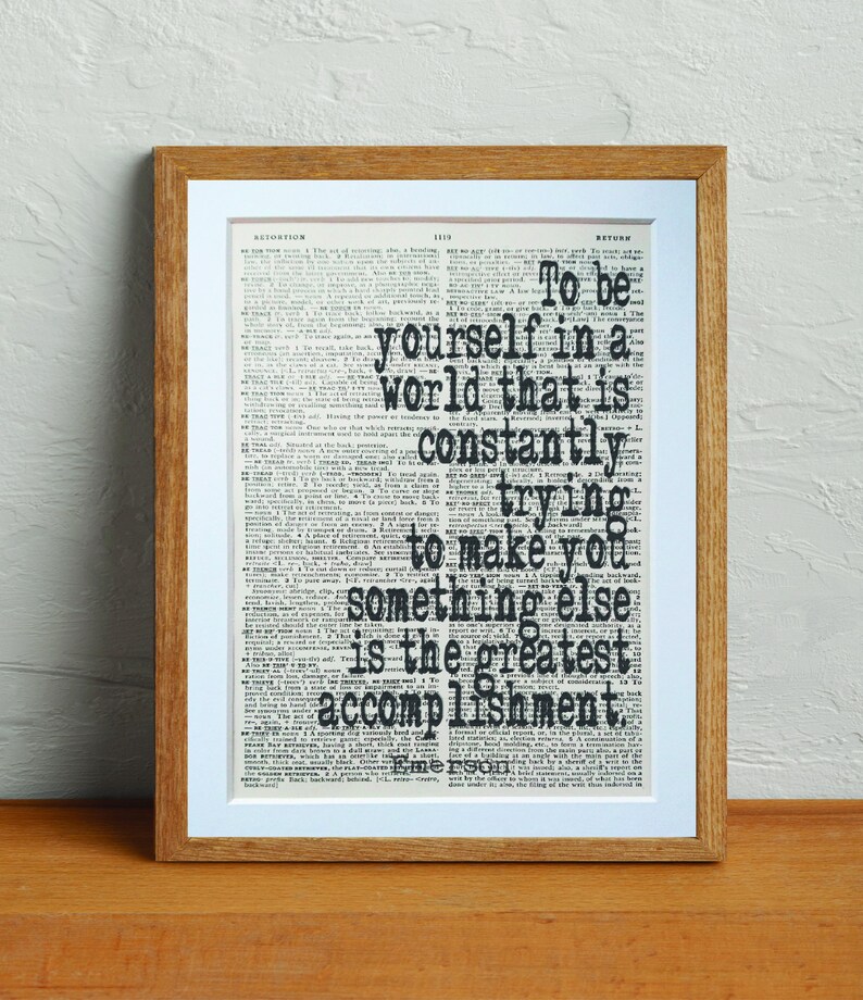 May include: A framed print with a quote on a vintage dictionary page. The quote reads, "To be yourself in a world that is constantly trying to make you something else is the greatest accomplishment." The frame is a light brown color.