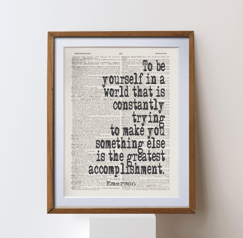 May include: A framed print featuring a quote by Emerson, "To be yourself in a world that is constantly trying to make you something else is the greatest accomplishment." The text is in black on a vintage book page.