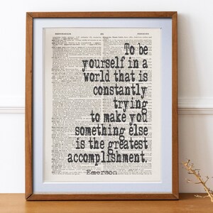 May include: A framed print featuring a quote by Emerson: "To be yourself in a world that is constantly trying to make you something else is the greatest accomplishment." The text is in black on a vintage dictionary page, framed in a brown wooden frame.
