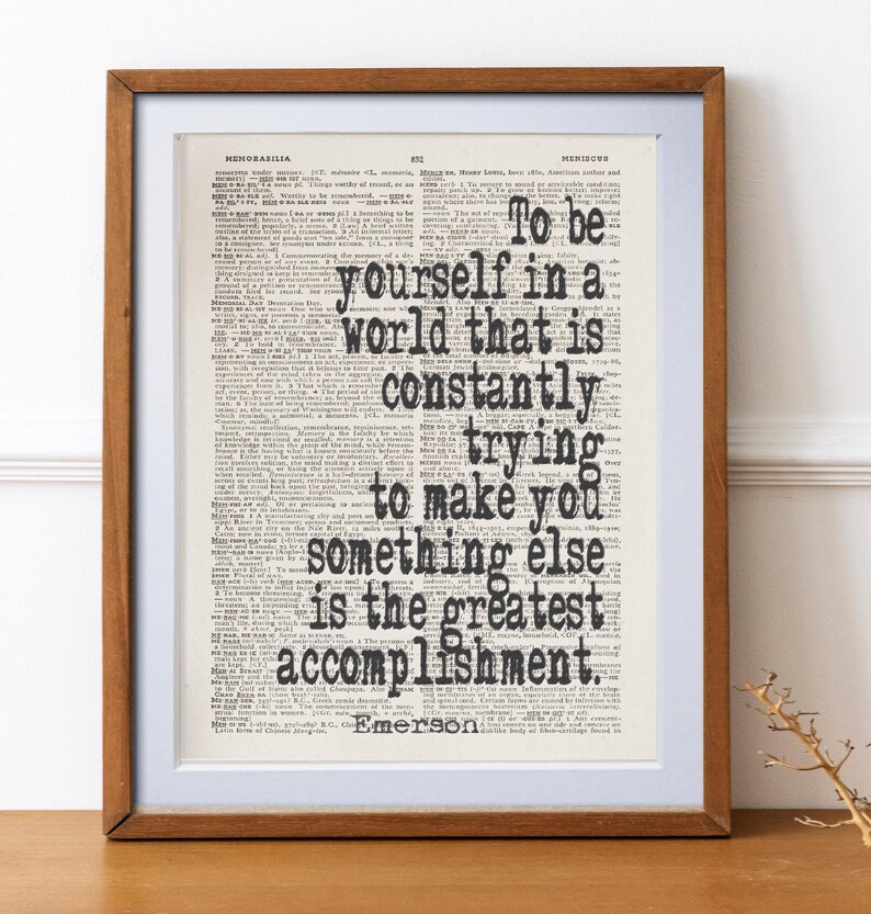 May include: A framed print featuring a quote by Emerson: "To be yourself in a world that is constantly trying to make you something else is the greatest accomplishment." The text is in black on a vintage dictionary page, framed in a brown wooden frame.