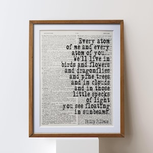 Könnte beinhalten: Gerahmter Druck mit einem Zitat von Phillip Pullman, vor einem Hintergrund aus einer alten Zeitung. Der schwarze Text lautet: „Every atom of me and every atom of you...“ Der Rahmen hat eine warme braune Farbe.