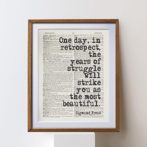 May include: Framed print featuring a quote by Sigmund Freud: "One day, in retrospect, the years of struggle will strike you as the most beautiful." The text is in black, set against a vintage dictionary page background, within a wooden frame.