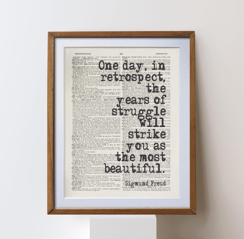May include: Framed print featuring a quote by Sigmund Freud: "One day, in retrospect, the years of struggle will strike you as the most beautiful." The text is in black, set against a vintage dictionary page background, within a wooden frame.