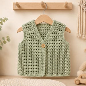 Peut inclure: Un gilet sans manches vert sauge en fil crocheté avec une encolure en V et une fermeture à un seul bouton en bois brun clair au centre. Le vêtement présente un motif à grille carrée répétitif sur toute la surface et est sur un cintre en bois.