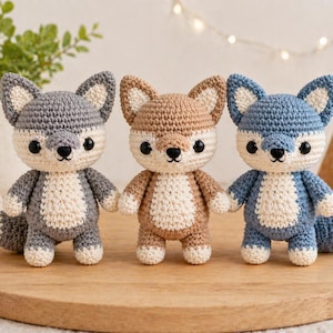 May include: Three handmade crochet fox toys in gray, brown, and blue. Each fox has a white belly, ears, and paws, with black safety eyes and a small black nose. The foxes are standing on a wooden surface.