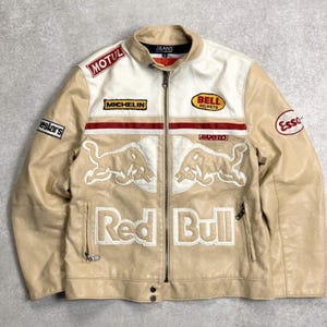 Op de afbeelding: Tan en witte leren jas met ritssluiting. De jas heeft patches met de logo's van "Motul", "Michelin", "Bell Helmets" en "Esso". De voorkant toont het "Red Bull" logo en een rode streep.