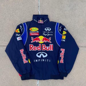 Puede incluir: Chaqueta de carreras Red Bull azul marino con múltiples logotipos de patrocinadores, incluidos Infiniti y Pepe Jeans. La chaqueta presenta un cuello alto, una cremallera completa y detalles en rojo y amarillo. El logotipo de Red Bull se muestra de forma destacada en la parte delantera.