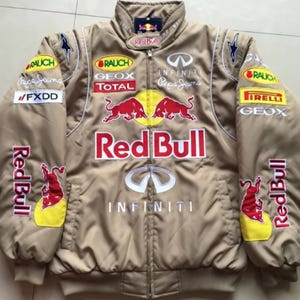 Puede incluir: Chaqueta de carreras Red Bull de color canela con múltiples logotipos de patrocinadores, incluidos Infiniti, Rauch y Total. La chaqueta tiene cuello alto, cierre de cremallera y varios parches y logotipos en rojo, amarillo y blanco.