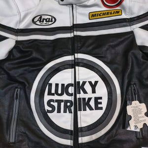 May include: Black and white leather motorcycle jacket with "LUCKY STRIKE" logo on the front. Features "MICHELIN" and "ARAI HELMET" patches. Includes a zip closure and a stand-up collar. Gray and white stripes accent the shoulders.