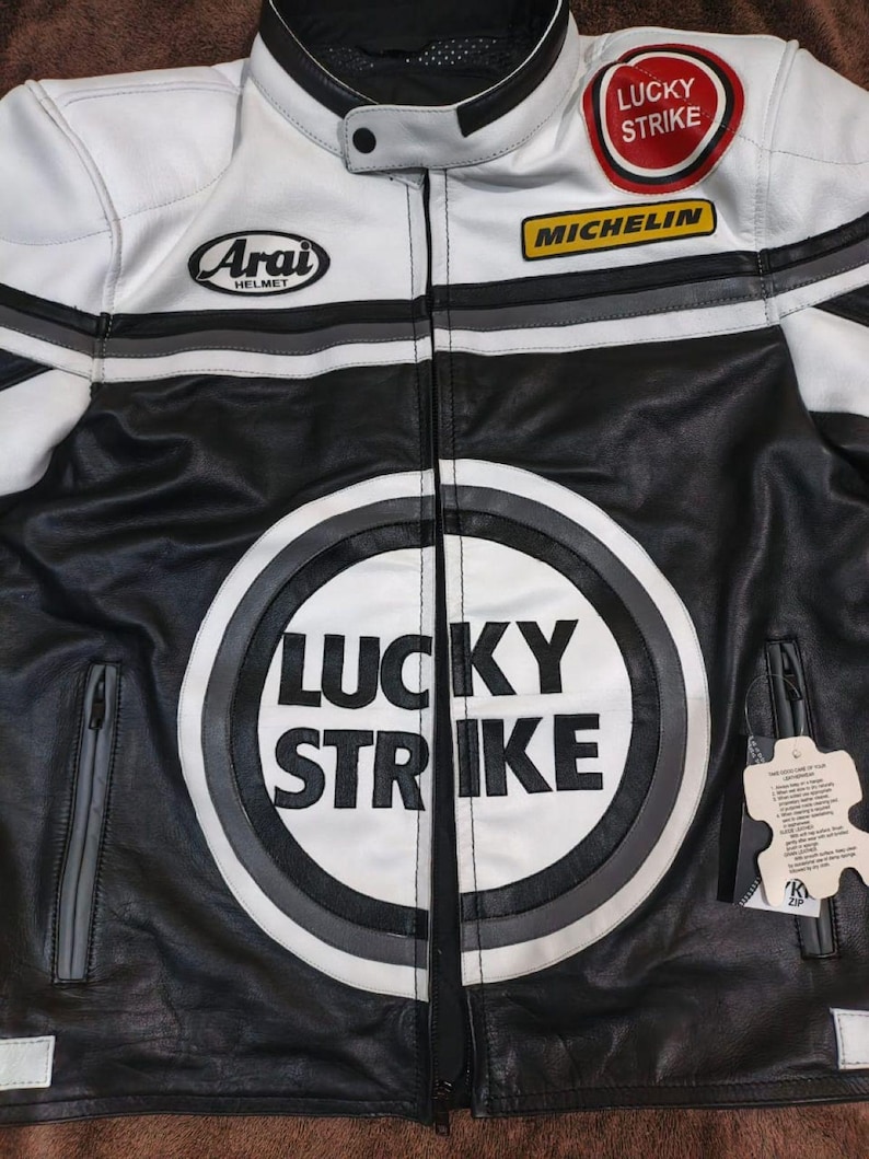 May include: Black and white leather motorcycle jacket with "LUCKY STRIKE" logo on the front. Features "MICHELIN" and "ARAI HELMET" patches. Includes a zip closure and a stand-up collar. Gray and white stripes accent the shoulders.