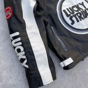 May include: Black leather jacket with white stripes and "LUCKY" printed on the sleeve. The jacket features a "LUCKY STRIKE" patch on the chest and sleeve. The jacket has a classic motorcycle style.