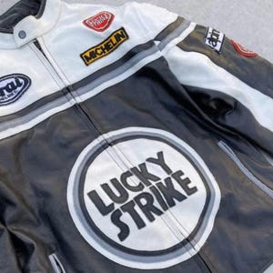 May include: Black and white leather motorcycle jacket with a zip-up front. The jacket features patches with the words "Lucky Strike" and "Michelin". Gray and white stripes accent the shoulders and sleeves.