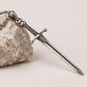 Könnte beinhalten: Ein schwertförmiger Anhänger aus silberfarbenem Metall an einer passenden Kugelkette. Das Schwert verfügt über ein detailliertes Heft mit Parierstange und strukturiertem Griff sowie ein oxidiertes Finish, das die Gravuren auf der Klinge betont.