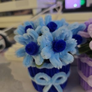 Op de afbeelding: Een handgemaakt bloemstuk van blauwe pijpenragers in een bijpassende blauwe pot met een strik. De bloemen zijn lichtblauw met donkerblauwe centra. De pot heeft een strikpatroon.