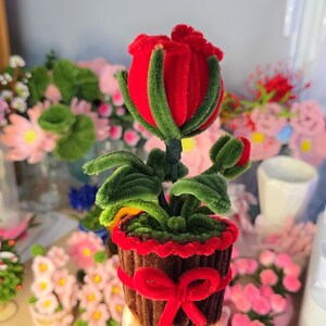 Puede incluir: Arreglo floral artesanal con una rosa roja brillante con detalles verdes, colocado en una maceta marrón adornada con un lazo rojo. La flor está hecha de un material suave y difuso, creando una estética única y caprichosa. Otros arreglos florales son visibles en el fondo.