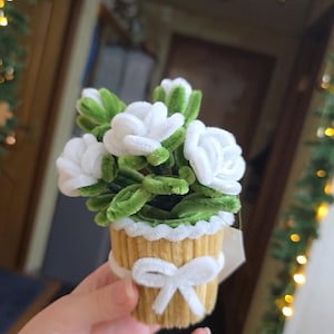 Op de afbeelding: Een handgemaakt bloemstuk met witte stoffen rozen en groene bladeren in een lichtbruine pot. De pot is versierd met een witte strik en een geschulpte rand.