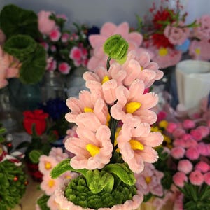 Può includere: Una composizione floreale artigianale con fiori rosa in scovolini e centri gialli, presentata in un contenitore giallo a forma di cupcake. La composizione è adornata con un fiocco rosa e accenti verdi. Altre composizioni floreali simili sono sullo sfondo.