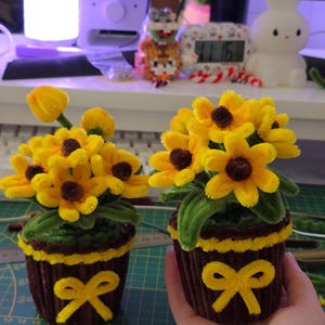 Op de afbeelding: Twee handgemaakte, miniatuur zonnebloemarrangementen in bruine cupcake-vormige potten. De zonnebloemen zijn geel met bruine centra en groene bladeren. De potten hebben gele strikken. De achtergrond omvat een computermonitor en een wit konijnenbeeldje.