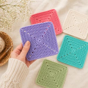 May include: A collection of five handmade crochet coasters in various colors: purple, pink, white, teal, and green. Each square coaster features a central starburst design and decorative corner cutouts. The coasters are displayed on a soft, neutral surface.