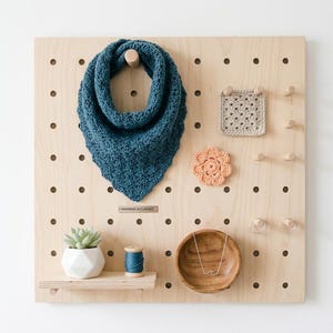 May include: A wooden pegboard showcases a blue knitted scarf, a beige crocheted square, and a peach-coloured crocheted flower. A small succulent in a white pot, a spool of blue thread, and a wooden bowl with a necklace are also featured. The pegboard has the text "HANDMADE IN FLORENCE".