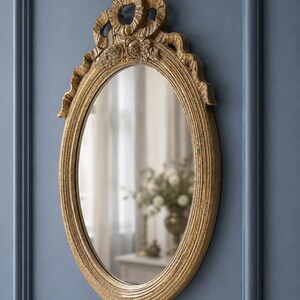 May include: Oval-shaped gold mirror with ornate detailing. The frame features a textured, ribbed design and decorative elements at the top, including floral accents and ribbon-like shapes. The mirror is mounted on a blue wall.