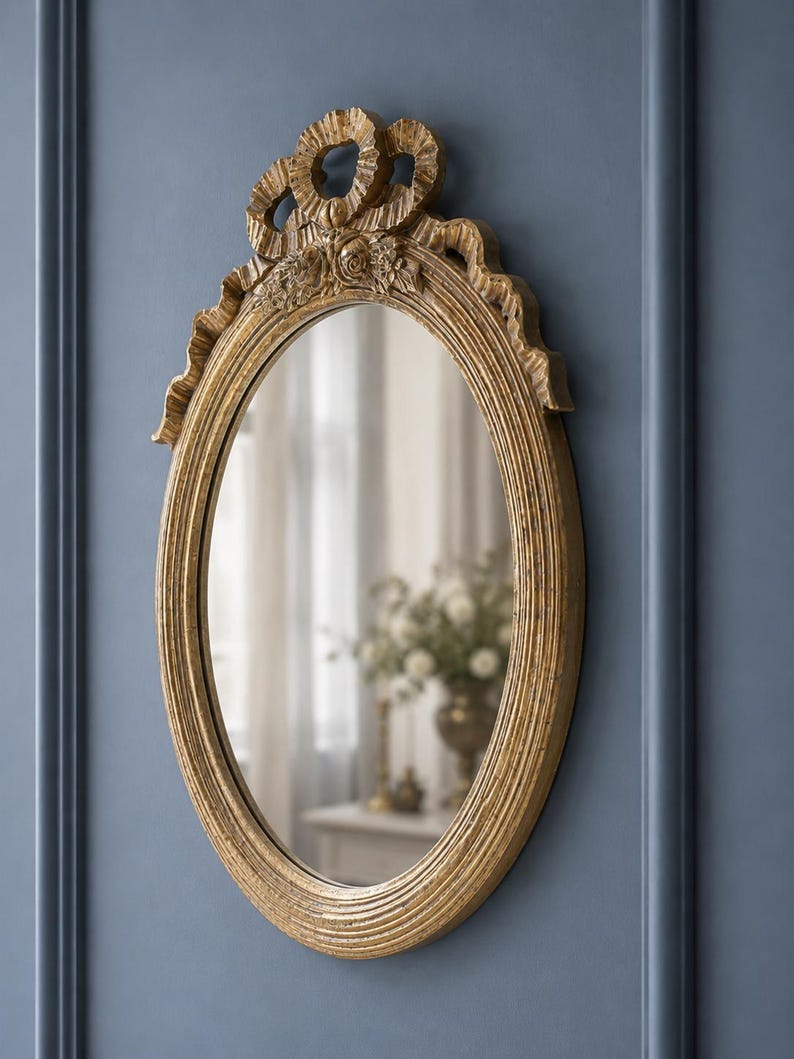 May include: Oval-shaped gold mirror with ornate detailing. The frame features a textured, ribbed design and decorative elements at the top, including floral accents and ribbon-like shapes. The mirror is mounted on a blue wall.