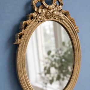 May include: Oval gold-colored mirror with ornate detailing. The frame features a ribbon and floral design at the top. The mirror is set against a blue background, reflecting a window and greenery.