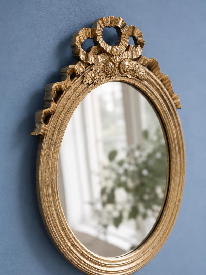 May include: Oval gold-colored mirror with ornate detailing. The frame features a ribbon and floral design at the top. The mirror is set against a blue background, reflecting a window and greenery.