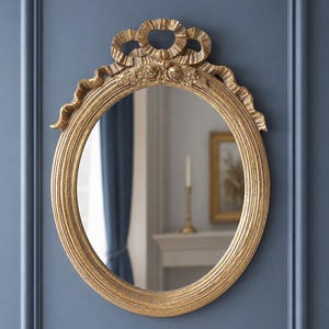 May include: Oval gold-colored mirror with a decorative ribbon and floral design at the top. The mirror has a ridged frame and is mounted on a blue wall. The reflection shows a glimpse of a room with a candle.